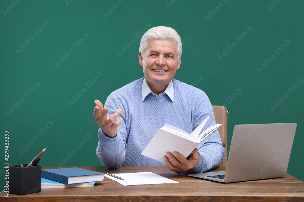 Portrait of senior teacher with laptop at table against green chalkboard Portrait of senior teacher with laptop at table against green chalkboard