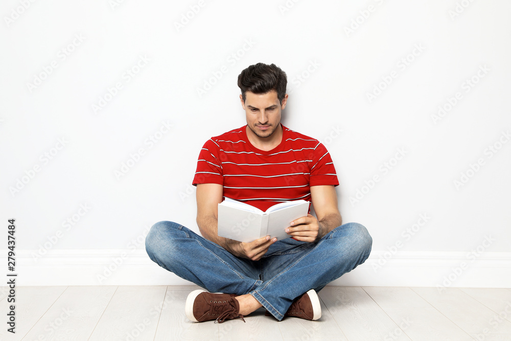 Handsome man reading book on floor near white wall