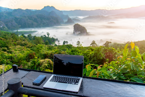 A laptop, mobile  on wooden table with sunrise and mountain fog background in morning. A start of new day. Freelance business concept. © theeraphong