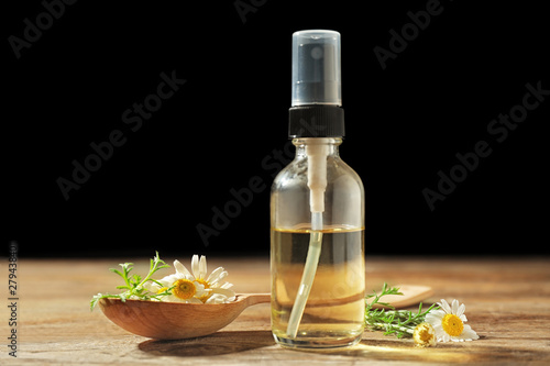 Bottle of chamomile essential oil and spoon with flowers on wooden table