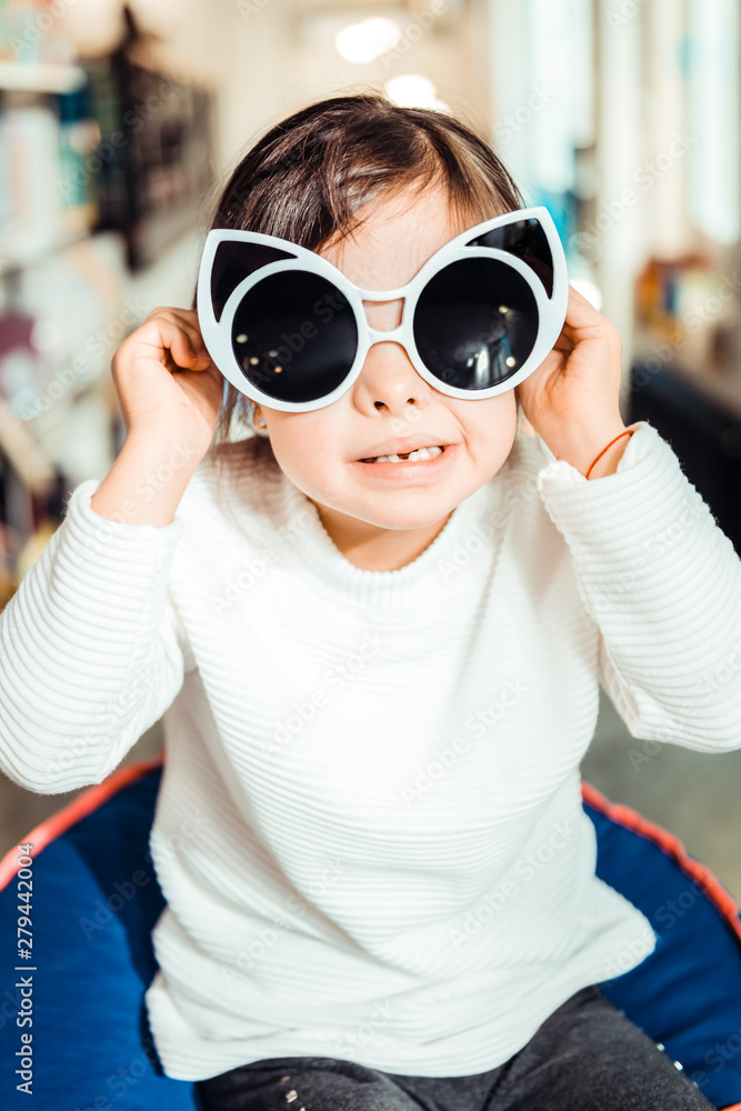 Dark-haired young girl in white sweater trying on giant sunglasses Dark-haired young girl in white sweater trying on giant sunglasses