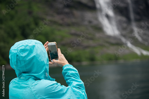 Girl in blue jacket taking photos of waterfall in Geiranger fjord, Norway