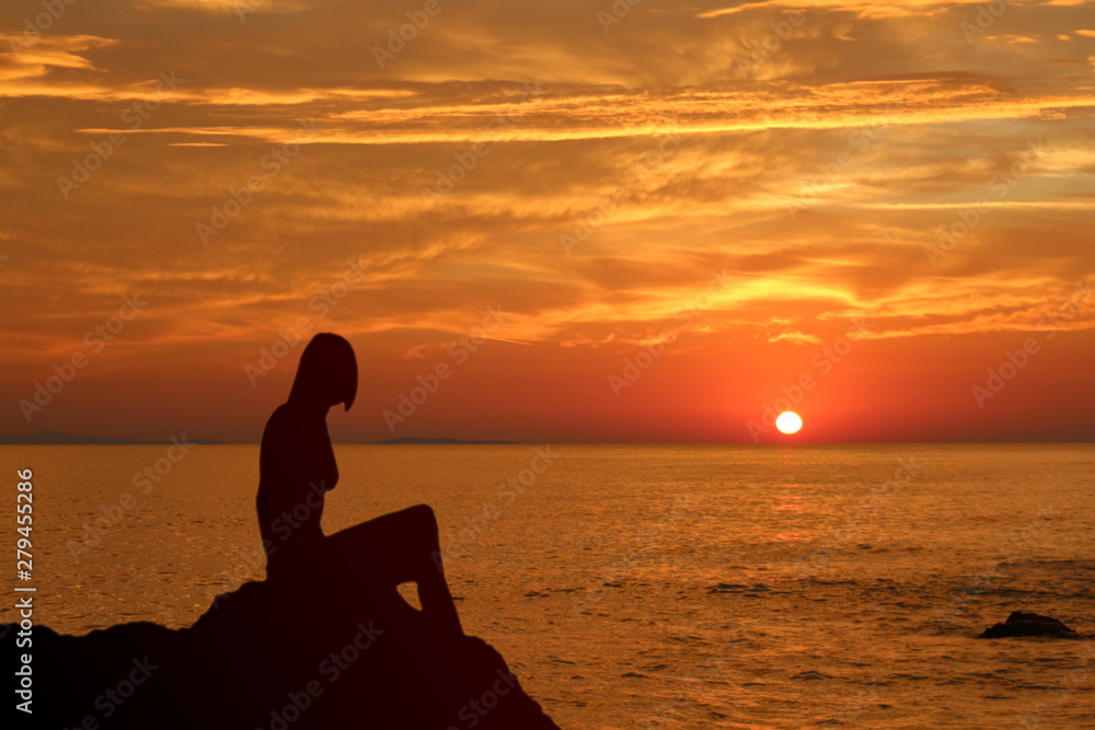 Silhouette of a young woman sitting on a rock against sunset at the sea