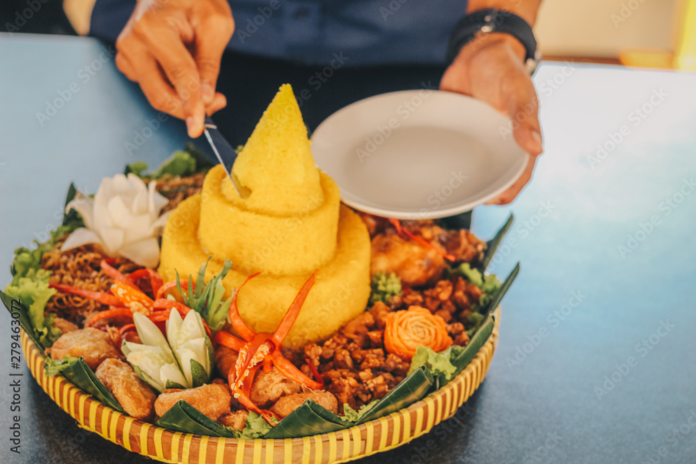 A hand cut Nasi Tumpeng Nusantara with knife for celebration. The elaborate  Indonesian rijsttafel of yellow rice with side dishes from several regional  cuisines in the country. Stock Photo | Adobe Stock
