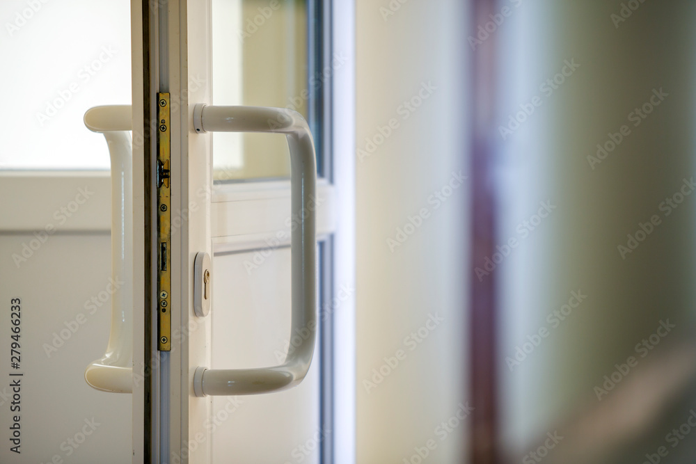 White plastic door detail with metal lock, handle and transparent glass on blurred interior background. Installation and design concept. White plastic door detail with metal lock, handle and transparent glass on blurred interior background. Installation and design concept.