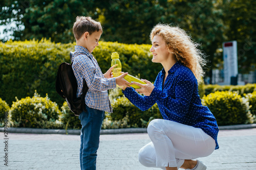 Blond curly caucasian mother giving school lunch and bottle with wather to her little son outdoors in sunny morning. Back to school concept. photo