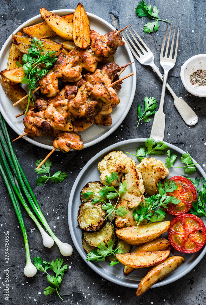 Fototapeta premium Meat skewers kebab with baked potatoes and roasted zucchini, tomatoes, cauliflower on a dark background, top view. Delicious tapas, appetizers in mediterranean style
