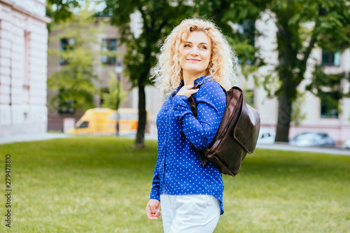 Adorable blond curly haired woman in blue shirt and brown leather bacpack, outdoor portrait. © Iryna