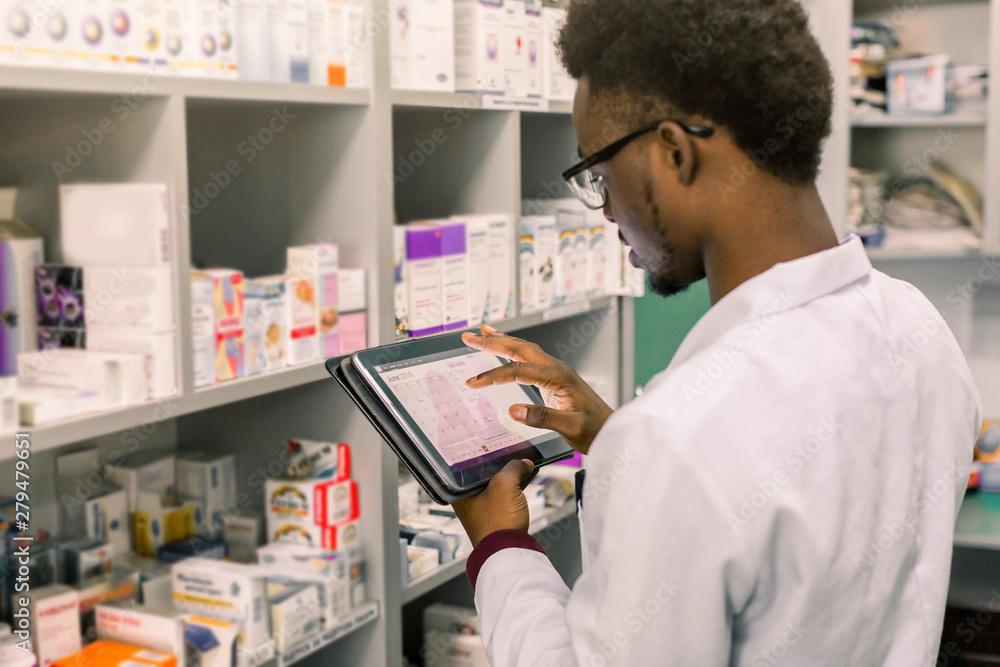 African American male pharmacist making notes on digital tablet in pharmacy, drug quality control African American male pharmacist making notes on digital tablet in pharmacy, drug quality control