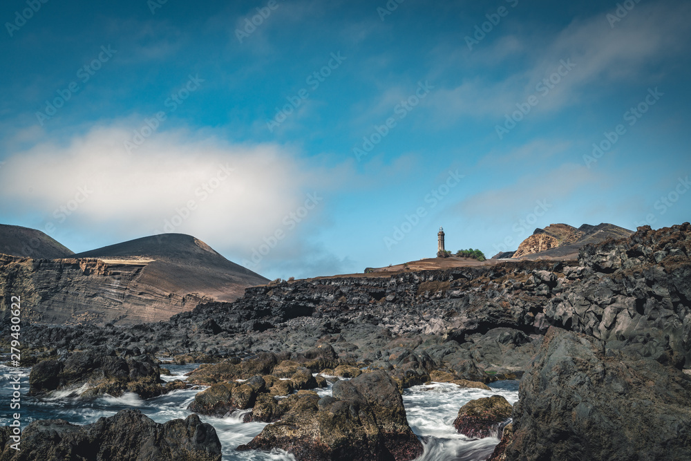 View over Capelinhos volcano, lighthouse of Ponta dos Capelinhos on western coast on Faial island, Azores, Portugal on a sunny day with blue sky and clouds and waves. Last volcano eruption was in 1957