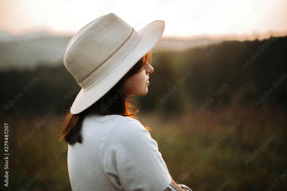 Stylish boho girl posing  in sunny light at atmospheric sunset in meadow. Happy hipster woman in poncho and hat enjoying traveling in evening mountains. Space for text