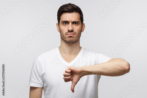Disgruntled grumpy dissatisfied annoyed unhappy young man showing thumb down as dislike gesture isolated on gray background