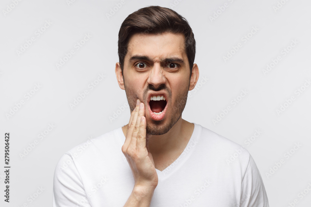 Toothache concept. Close-up of young man isolated on gray background touching his face with expression of horrible suffer from health problem and aching tooth Toothache concept. Close-up of young man isolated on gray background touching his face with expression of horrible suffer from health problem and aching tooth