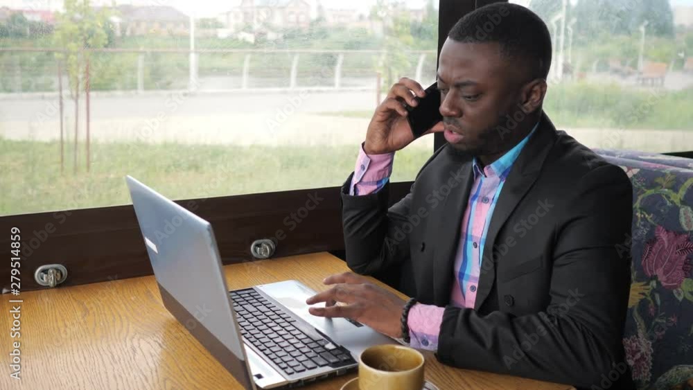 Afro american businessman working on laptop and talking mobile phone in cafe. He has a cup of coffee. Black man calling mobile phone and looking documents on computer. He wears shirt and suit jacket. Afro american businessman working on laptop and talking mobile phone in cafe. He has a cup of coffee. Black man calling mobile phone and looking documents on computer. He wears shirt and suit jacket.