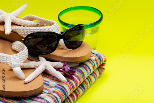 Colorful Striped Beach Towel, Women's Causal Flip Flops, Blue Rim Glass with Water and Black Sunglasses on yellow background as Summer Vacation Theme.