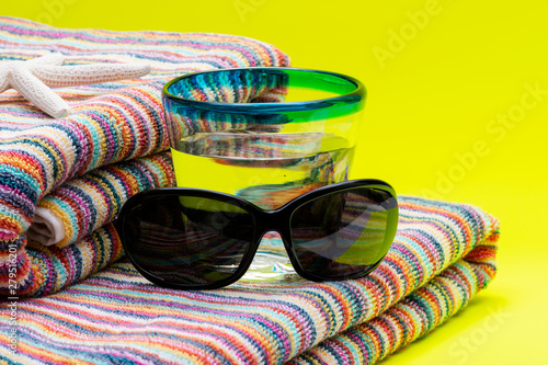 Folded Colorful Striped Organic Cotton Beach Towels, Blue Rim Glass with Water and Black Sunglasses on yellow background as Summer Vacation Concept.