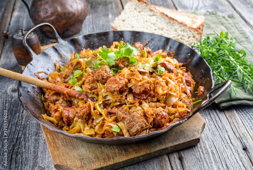 Traditional Polish kraut stew bigos with sausage, meat and mushrooms as closeup in a wrought-iron pan on an old wooden table - obrazy, fototapety, plakaty 