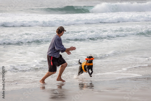 Fotografi Man playing with his dog on the beach with fetching toy while waves are crashing