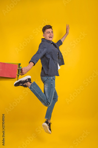Impressed young man with colorful shopping bags in hands with open mouth, happy after successful shoping, isolated over yellow background