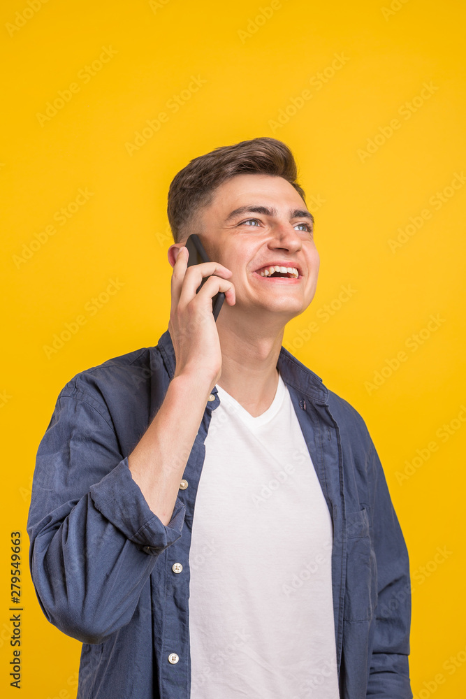 Handsome young man using his phone with smile while standing against yellow background.Young adult man talking on the phone scared in shock with a surprise face, afraid and excited with fear expressio Handsome young man using his phone with smile while standing against yellow background.Young adult man talking on the phone scared in shock with a surprise face, afraid and excited with fear expressio