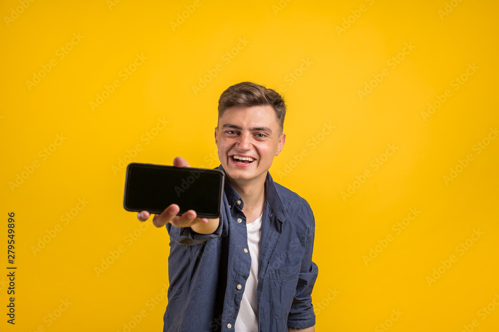 Attractive young man with smartphone on color yellow background. Close up portrait of a satisfied young man in t-shirt showing blank screen mobile phone isolated over yellow background Attractive young man with smartphone on color yellow background. Close up portrait of a satisfied young man in t-shirt showing blank screen mobile phone isolated over yellow background