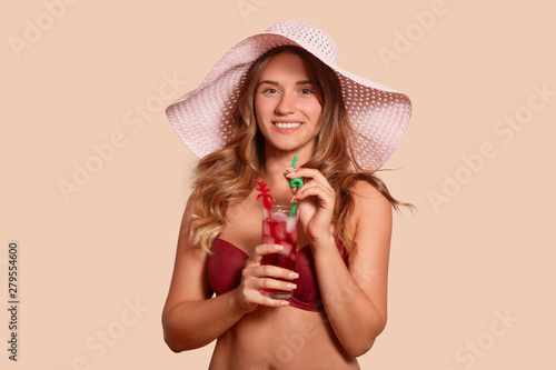Magnetic satisfied young woman wearing hat and swimming suit, holding glass of cocktail, standing isolated over beige background in studio, looking directly at camera. Vacation time concept.
