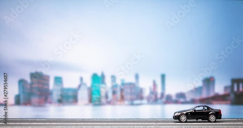 Time lapse of a black car figurine moving from right to left on a highroad model with a city image in the background, passing twice.