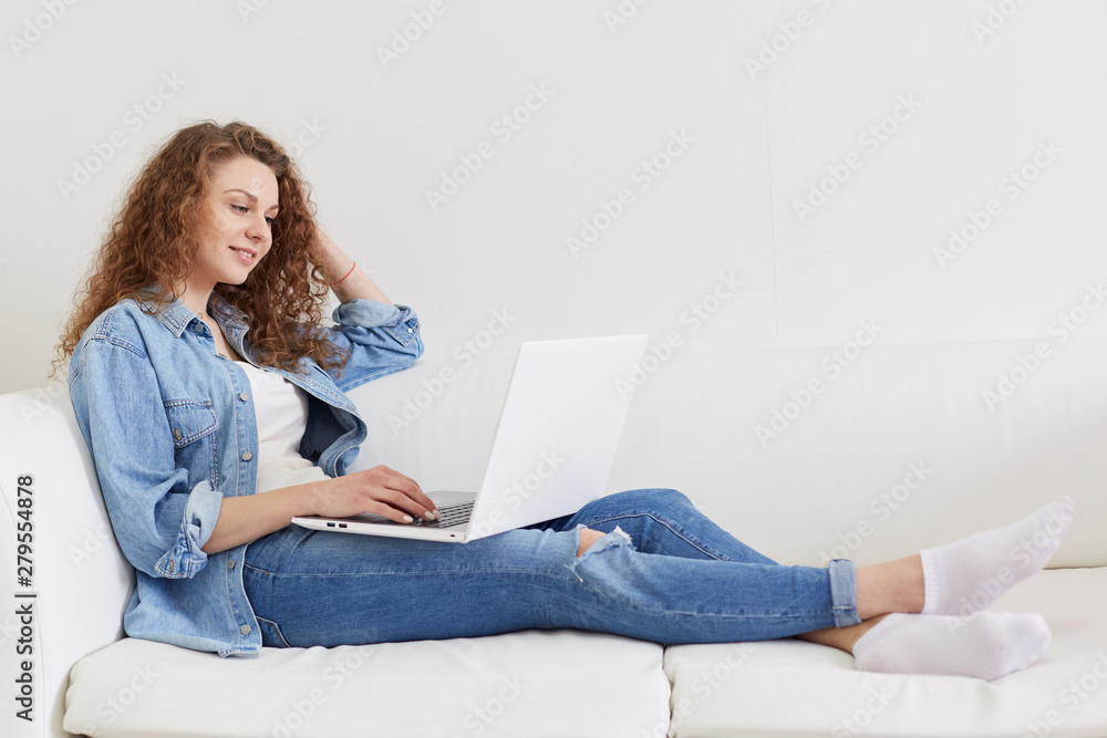 Horizontal shot of good looking curly haired young model sitting on white sofa, having laptop, watching videos, applying information, wearing jeans, white t shirt, socks and jacket. Tech concept. Horizontal shot of good looking curly haired young model sitting on white sofa, having laptop, watching videos, applying information, wearing jeans, white t shirt, socks and jacket. Tech concept.