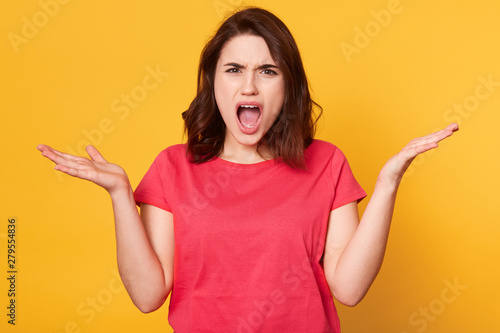 Close up portrait of angry disappointed brunette shouting with dissatisfaction, opening her mouth widely, looking directly at camera, raising her hands, standing isolated over yellow background.
