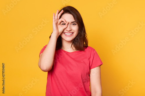 Image of peaceful cheerful young model posing isolated over bright yellow background in studio, putting her fingers to face, making gesture OK, smiling sincerely, wearing red casual t shirt.