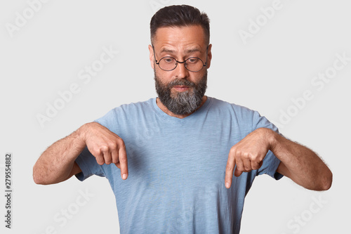 Studio shot of handsome charismatic model, wearing blue casual t shirt and round eyeglasses, having beard, making gesture, showing direction with his forefingers. Copyspace for advertisement.