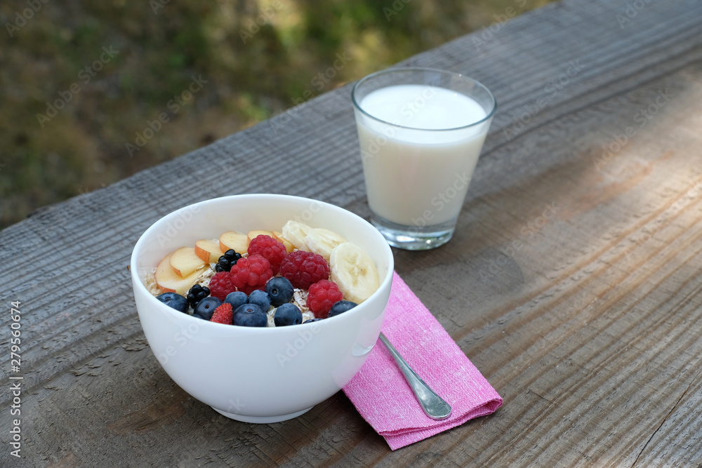 Close up a white bowl of rolled oats served with milk, slices of bananas, fresh apple, wild strawberry, blue berries, red raspberries and blackberries. Healthy breakfast outside in summer. Close up a white bowl of rolled oats served with milk, slices of bananas, fresh apple, wild strawberry, blue berries, red raspberries and blackberries. Healthy breakfast outside in summer.
