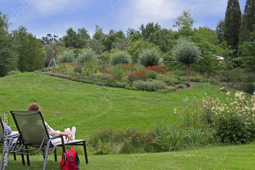 Sommerlicher Park mit grüner Wiese und farbenfrohen Blumenbeeten, Menschen im Liegestuhl genießen das sonnige Wetter