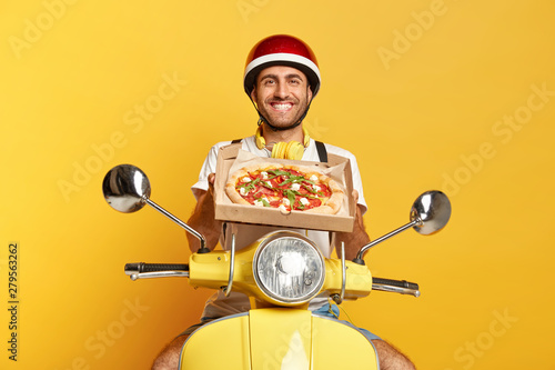 Satisfied motorcyclist delivers tasty fresh baked pizza, wishes good appetite for customers, holds cardboard box, wears protective helmet, sits on fast motorbike, isolated on yellow studio wall