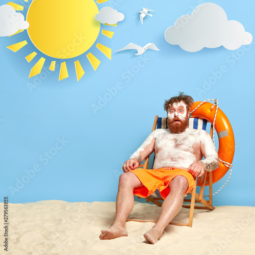 Shocked man has sunscreen cream on body to protect skin from uv rays, has summer vacation, burnt skin during sunbathinng, sits on comfortable beach chair, enjoys good rest during hot sunny day. photo