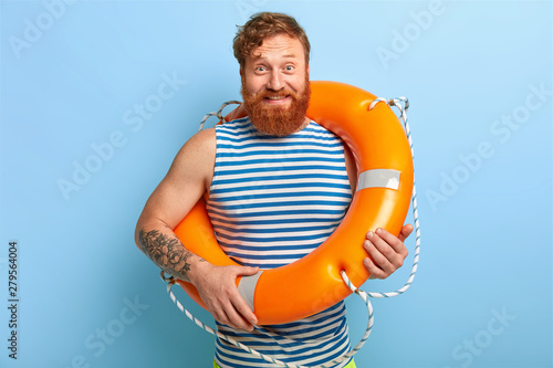 Glad man rescuer poses with orange lifebuoy, wears striped sailor vest, has red hair and beard, rests during summer holiday, has tattoo on arm, isolated on blue background, ready for swimming