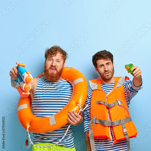 Friends with swimming equipment, use water pistols for battle, wear striped sailor jumpers, have fun at beach during good summer weather, pose against blue background, enjoy vacation at seaside