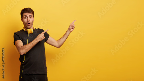 Shot of shocked youngster reacts on cool promo, points at blank space, recognizes something incredible, wears black clothes with headphones, isolated on yellow background. Unexpected scene happened © wayhome.studio 