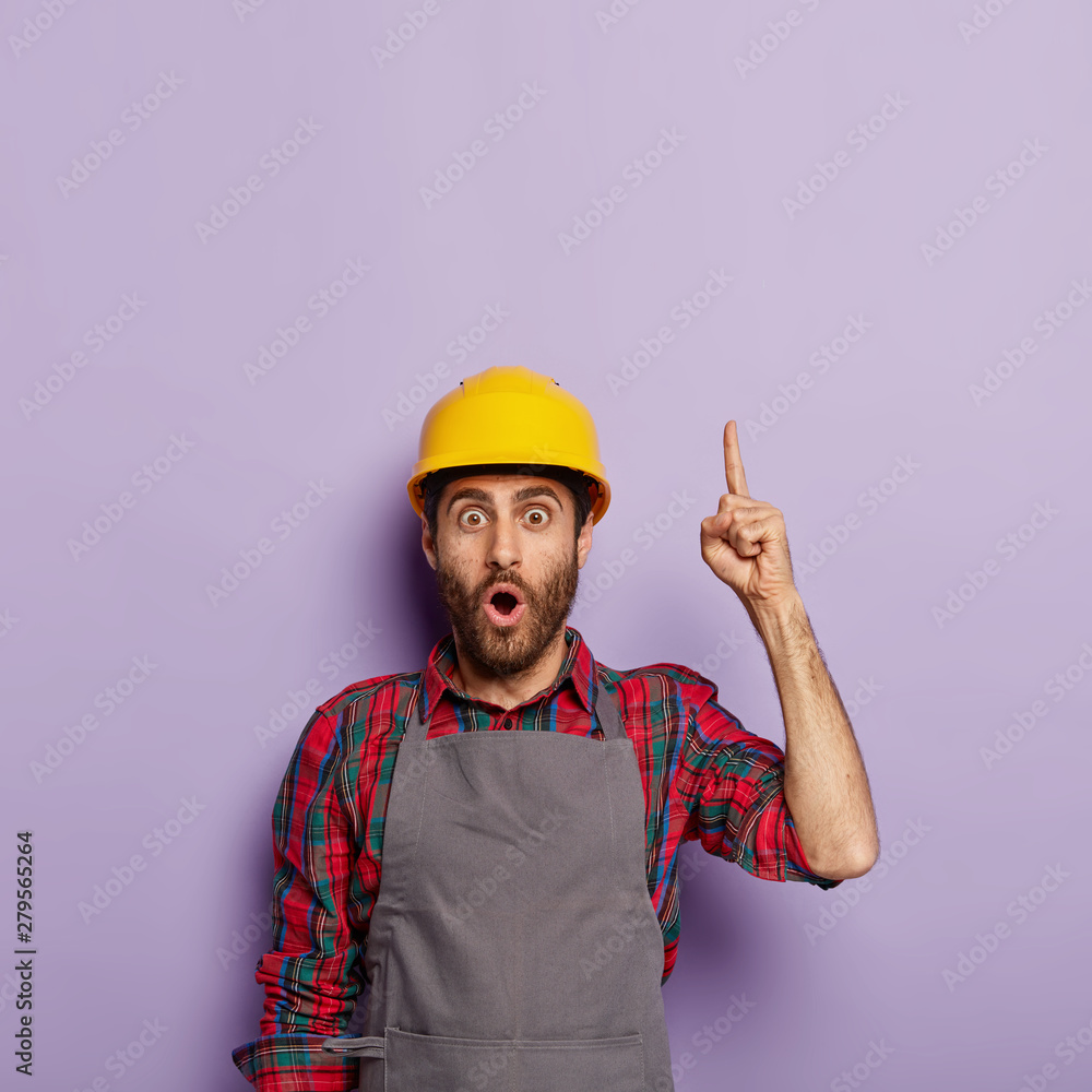 Shocked male industrial worker wears yellow hardhat, apron, points upwards, shows blank space above, isolated on purple background. Mechanical worker or repairman in uniform busy with hard work. Shocked male industrial worker wears yellow hardhat, apron, points upwards, shows blank space above, isolated on purple background. Mechanical worker or repairman in uniform busy with hard work.
