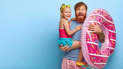 Image of happy redhaired father and daughter have glad expressions, pose with swimming equipment, ready for nice summer rest at seaside, isolated on blue wall with blank space. Holidays together