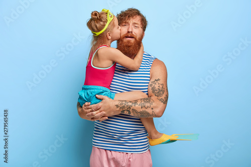 Small active girl wears goggles and flippers for swimming, kisses in cheek daddy with love, being at sea on summer vacation. Tired father carries daughter on hands, receives warm hug and kiss