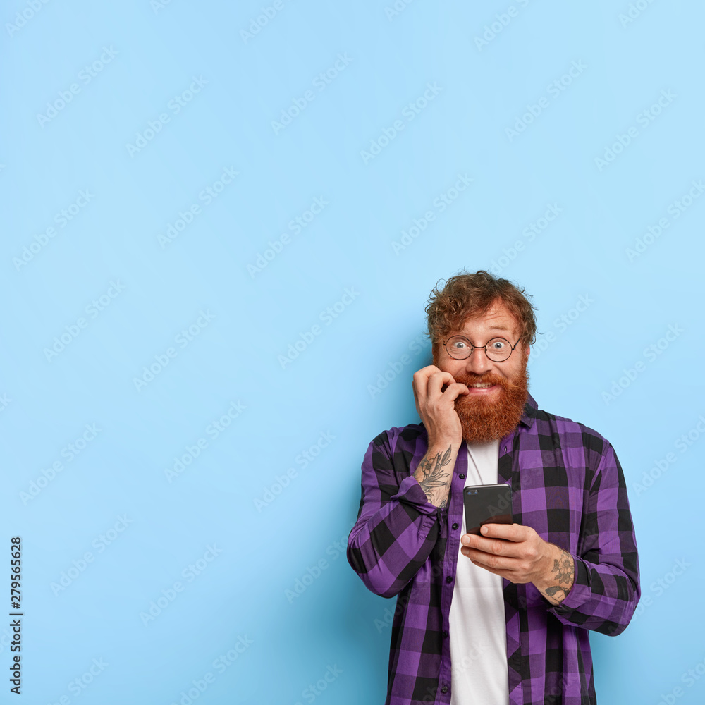 Fotografie Funny red haired man with nervous expression, bites finger nails, browses networ