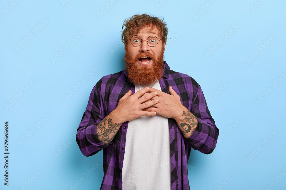 Φωτογραφία Excited redhead man cannot believe in something incredible, touches chest, looks with great amazement, smiles from positive emotions, wears round glasses, isolated on blue wall