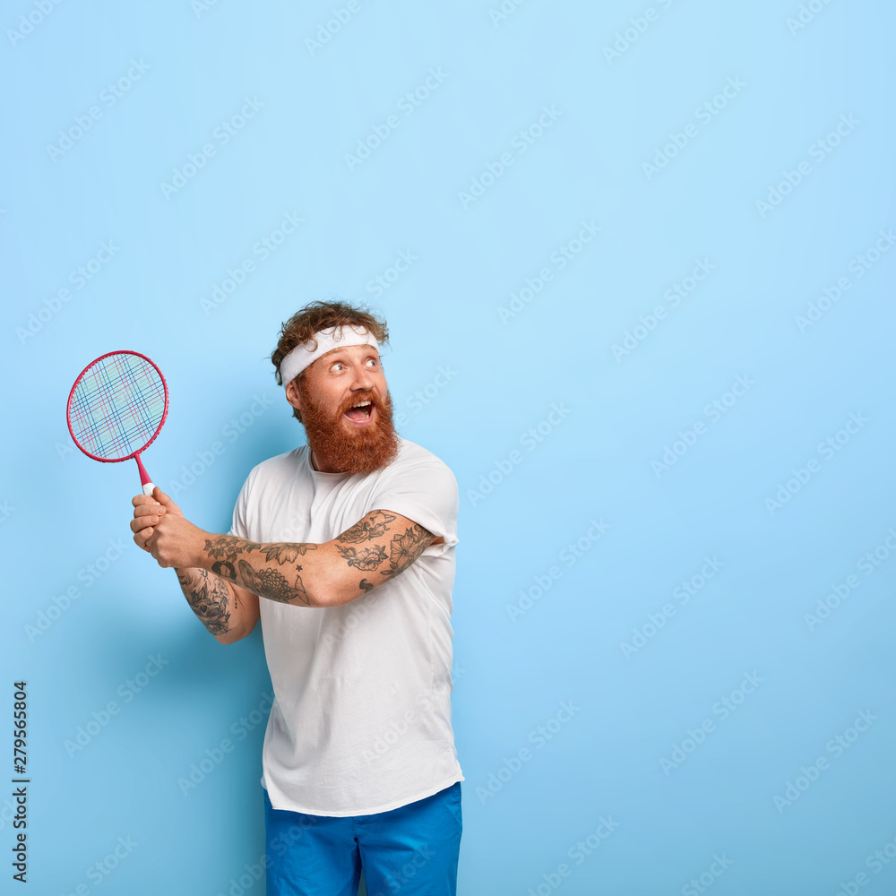 Cheerful active player focused happily into distance, holds racket and plays badminton, has attentive look, ready to serve, dressed casually, stands alone against blue studio wall with empty space Cheerful active player focused happily into distance, holds racket and plays badminton, has attentive look, ready to serve, dressed casually, stands alone against blue studio wall with empty space
