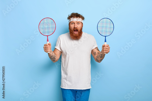 Outraged sporty man holds two badminton rackets, angry friend didnt come on match, needs companion for playing, screams with annoyance, wears white headband, sport outfit. Hobby and game concept © wayhome.studio 