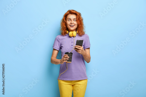 Photo of surprised cheerful woman has ginger hair, reacts at received message on smartphone, holds paper cup of coffee, wears purple t shirt and yellow trousers, checks mailbox. Technology concept © wayhome.studio 