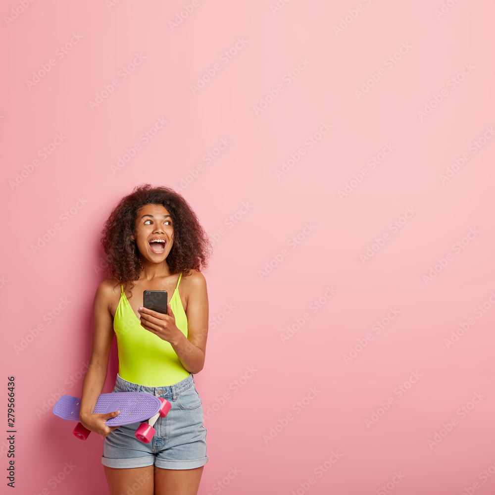 Overjoyed lovely teenager with bushy Afro hair, holds mobile phone, surfes social networks online during break after skateboarding, holds penny board, wears casual outfit, gazes on right side Overjoyed lovely teenager with bushy Afro hair, holds mobile phone, surfes social networks online during break after skateboarding, holds penny board, wears casual outfit, gazes on right side