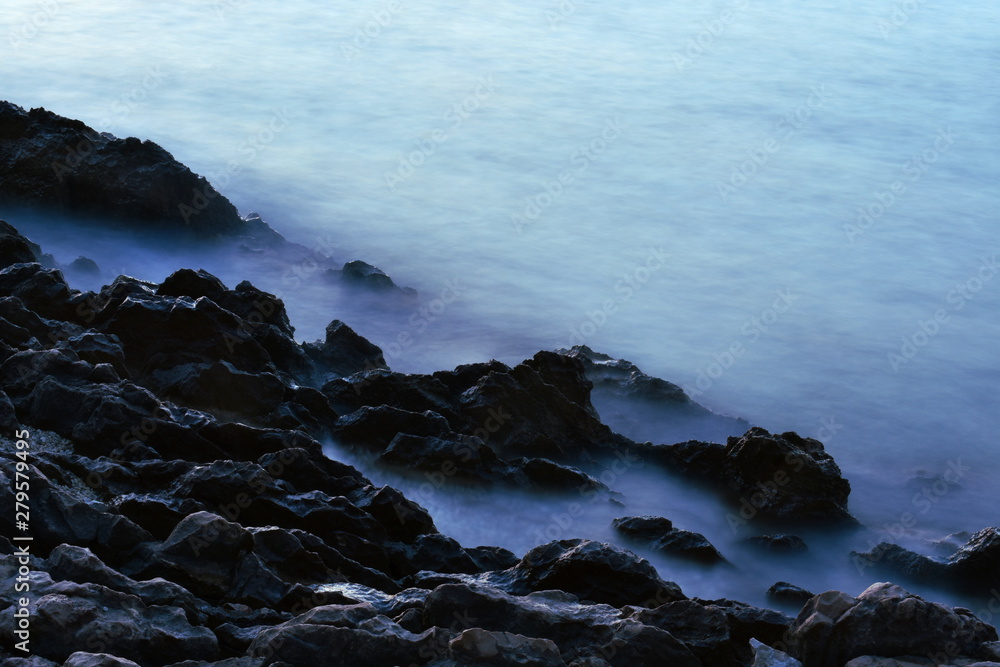 Rough rocky seashore with soft misty cloudy blue water. Horizontal, long exposure.