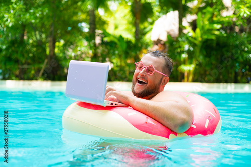 laughing Fat funny man in pink inflatable circle in pink glasses works on a laptop in a swimming pool