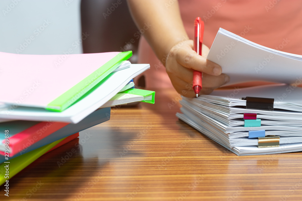 Teacher hand holding red pen for checking students homework assignment and pile of unfinished paperwork stacked in archive with colorful plastic slide binder bar beside. Business and education concept Teacher hand holding red pen for checking students homework assignment and pile of unfinished paperwork stacked in archive with colorful plastic slide binder bar beside. Business and education concept